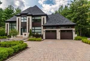 Modern luxury house with dark wood garage doors, stone exterior, and contemporary architectural design surrounded by landscaped greenery and a spacious paved driveway.
