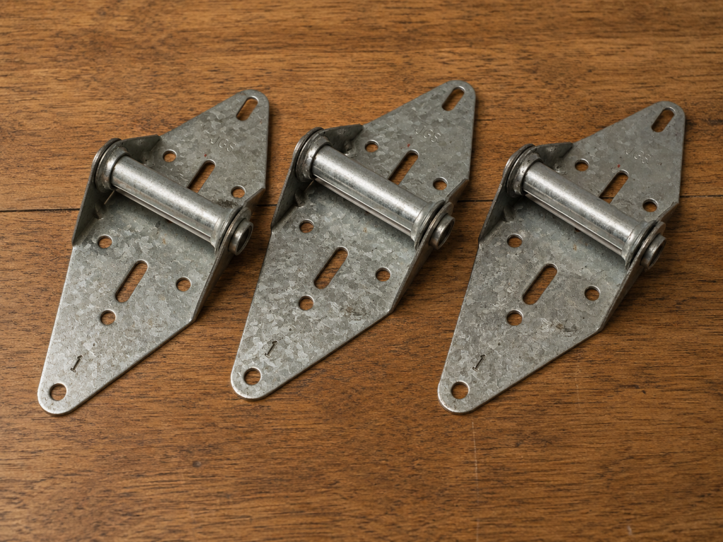 Three galvanized steel garage door hinges arranged on a wooden workbench, showing common replacement hardware used for garage door panel support, alignment, and smooth operation.